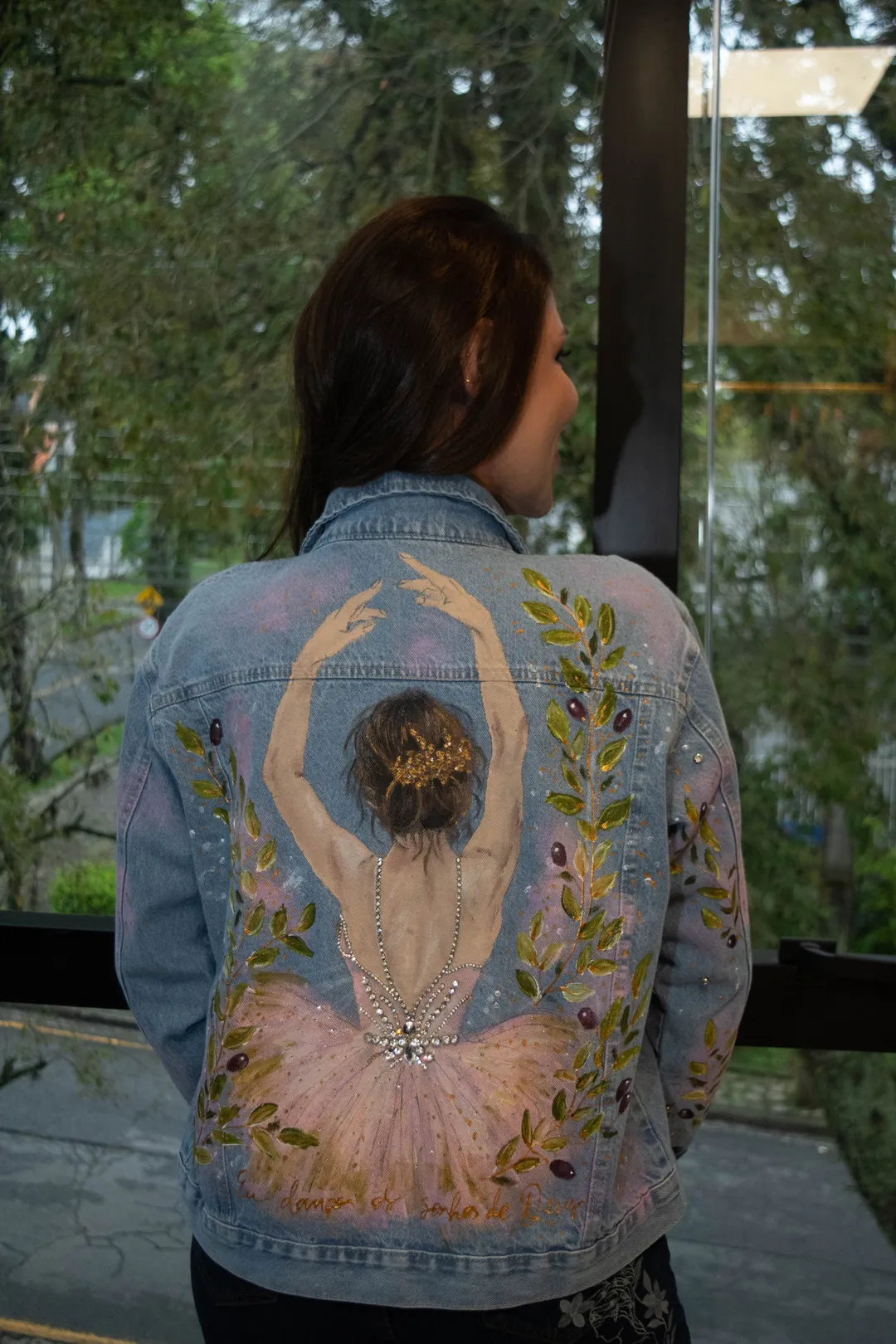 Mulher de costas vestindo jaqueta jeans com pintura de bailarina de saia rosada, braços erguidos e ramos verdes.