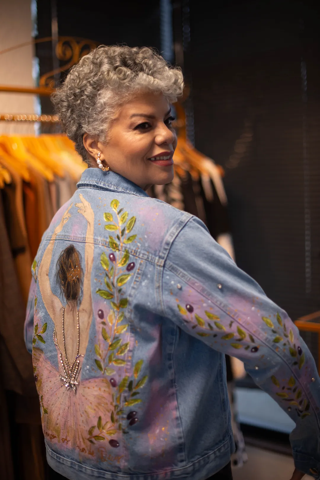 Mulher sorrindo por cima do ombro, vestindo jaqueta jeans R U T H. com pintura de bailarina e ramos de oliveira nas costas e mangas.
