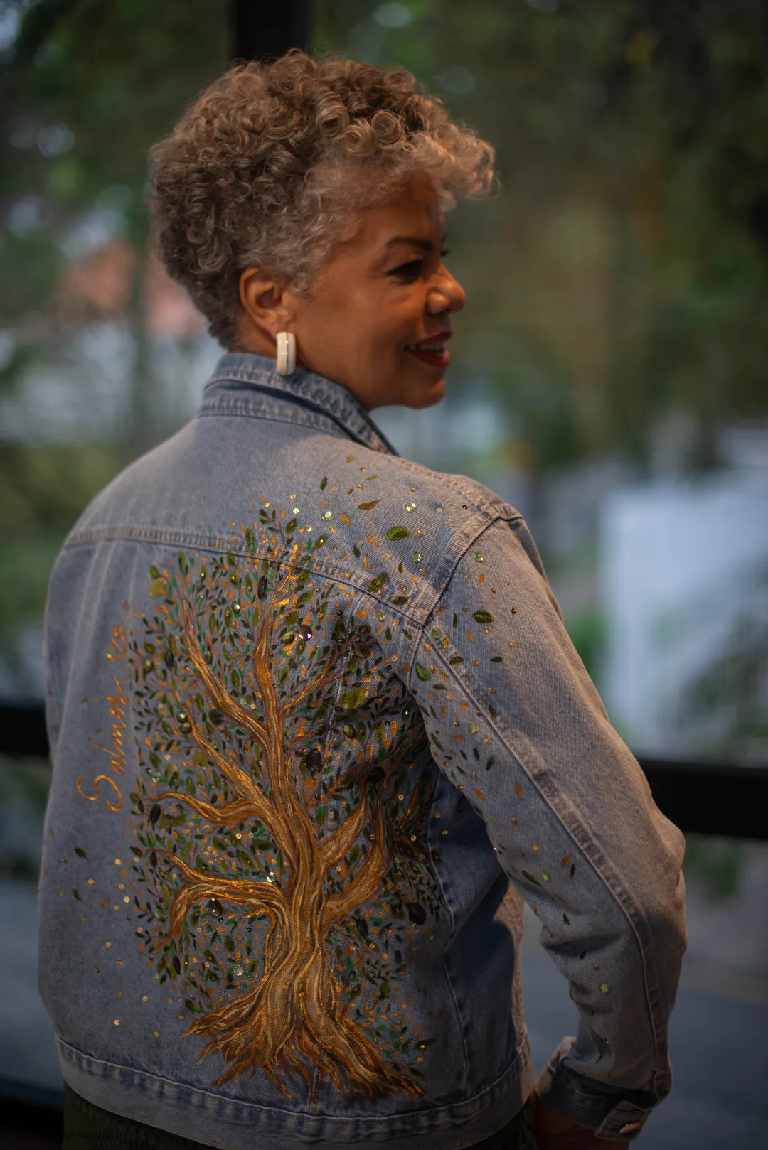 Mulher de costas vestindo jaqueta jeans com pintura de árvore de tronco dourado e folhagem com pedrarias.