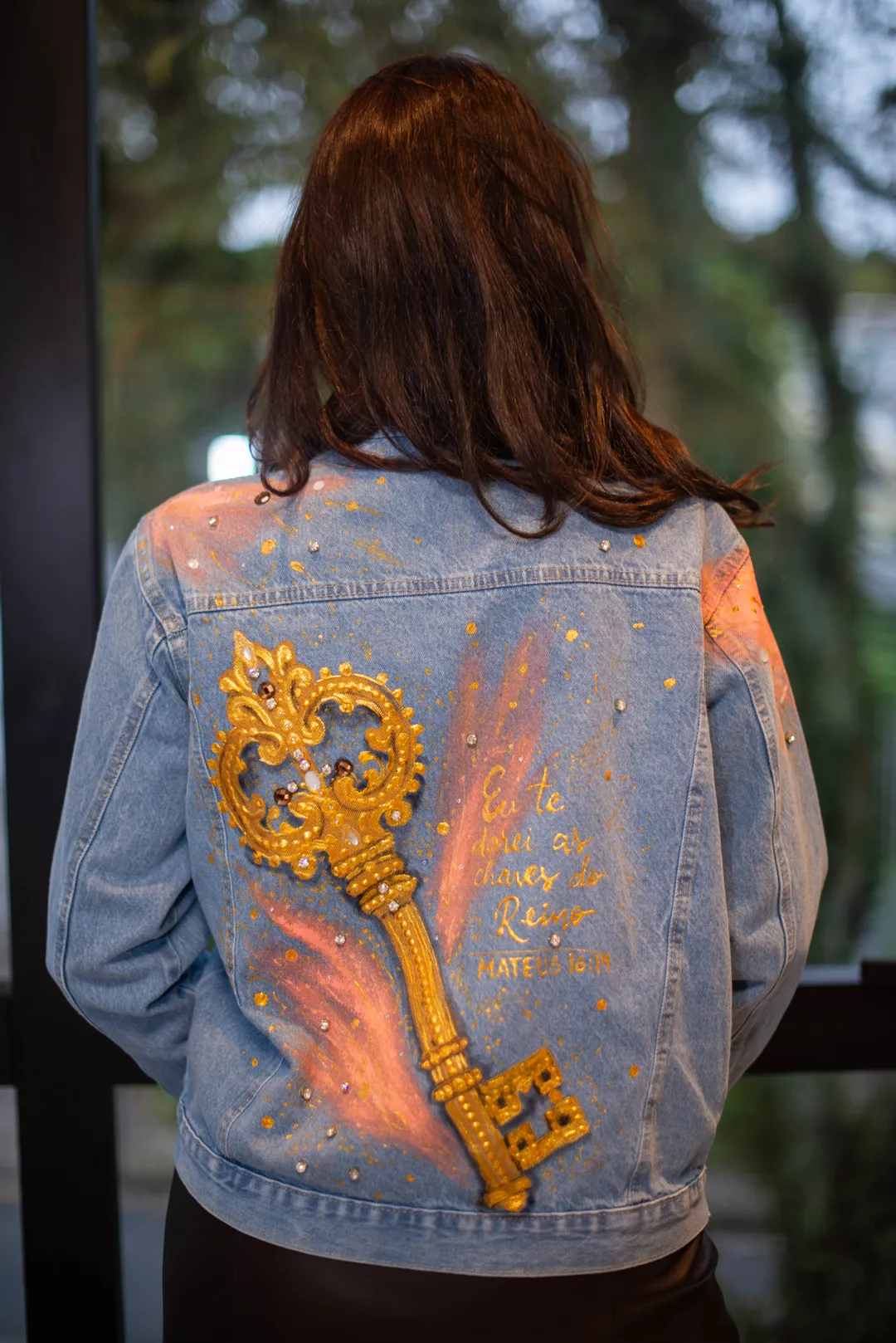 Mulher de costas vestindo jaqueta jeans com chave dourada em relevo, asas em tons pêssego e inscrição manuscrita.