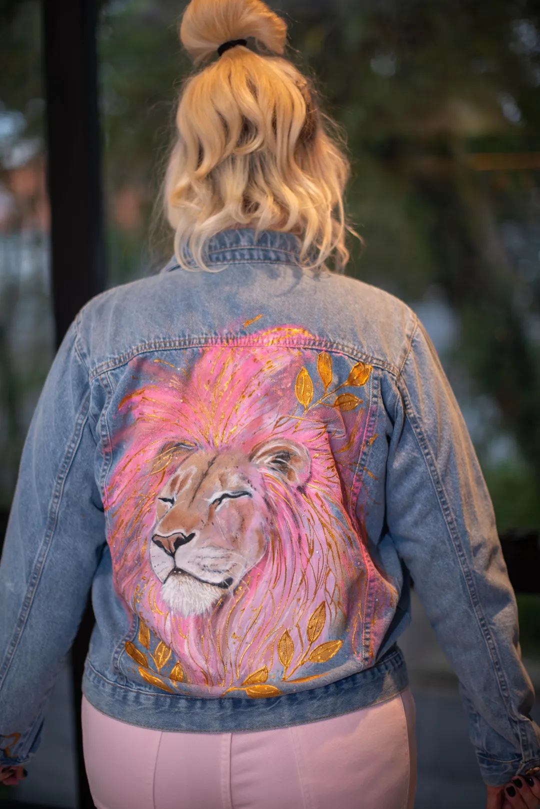 Mulher de costas vestindo jaqueta jeans com pintura de leão em tons rosados e folhas douradas.