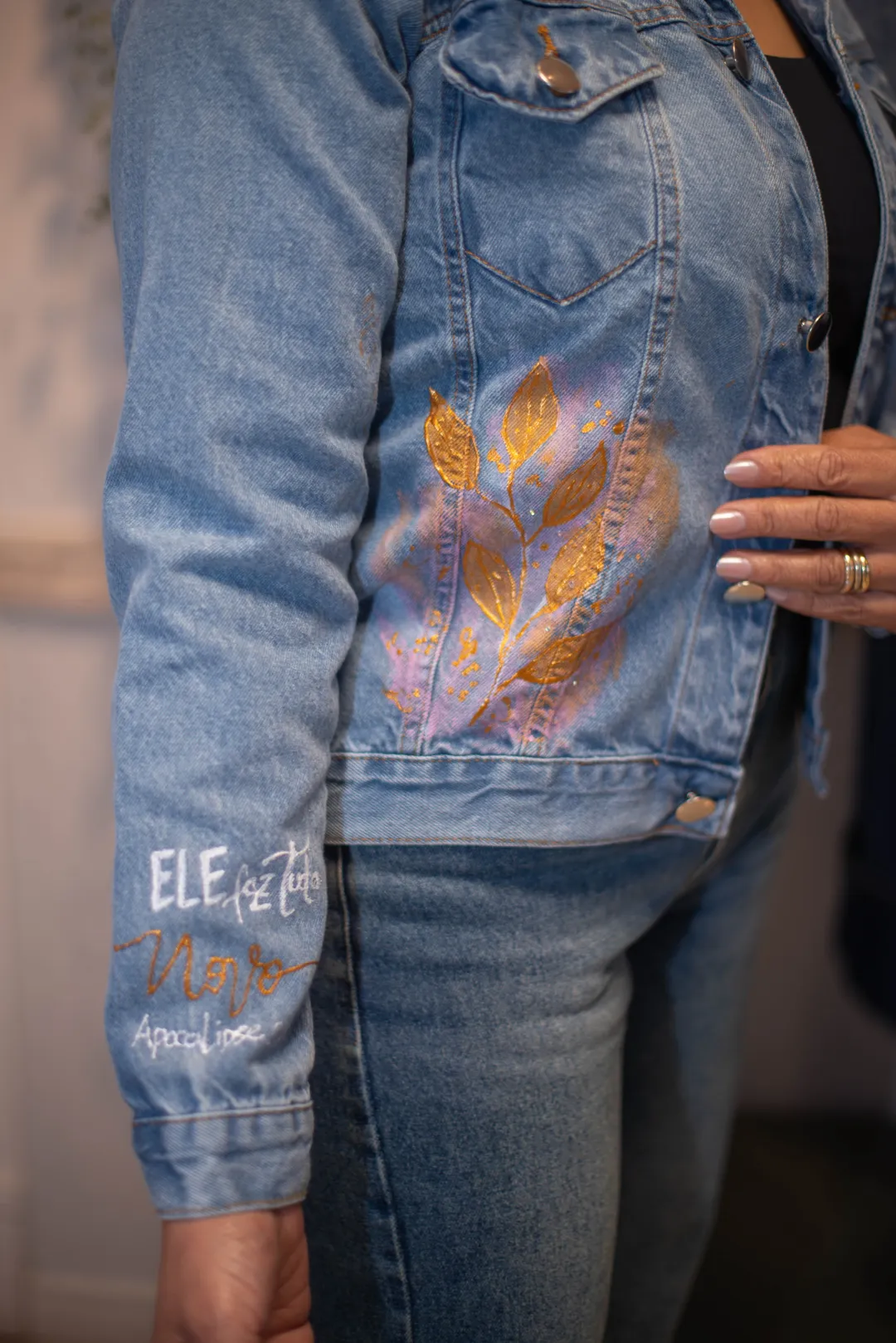 Manga de jaqueta jeans com inscrição pintada à mão, com referência ao livro de Apocalipse, ao lado de folhas douradas.